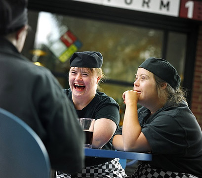 Tre camerieri scherzano seduti intorno ad un tavolino, davanti ad una bibita, in una stazione. Kate, una di loro, ha la sindrome di Down. © Richard Bailey